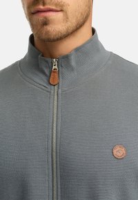 Parte inferior de la cara y cuello de un hombre que lleva una chaqueta gris de cremallera con textura, con un tirador de cremallera de cuero marrón y un parche redondo marrón con un logo en el pecho.
