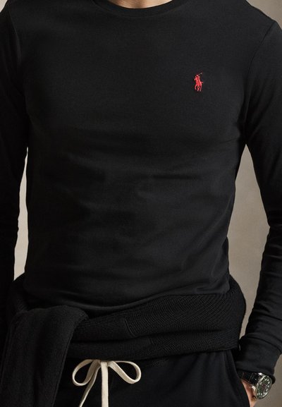 Mężczyzna ubrany w czarną koszulę z długim rękawem z czerwonym logo gracza polo, czarne spodnie z białym ściągaczem i czarny sweter związany wokół talii.