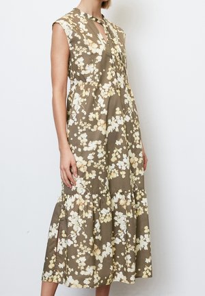 Femme portant une robe midi sans manches vert olive à imprimé floral blanc et jaune avec un ourlet à volants, debout devant un fond blanc uni.