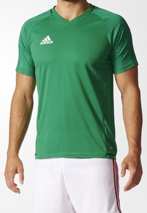 Mężczyzna w zielonej sportowej koszulce Adidas z krótkim rękawem i dekoltem w serek oraz białych szortach z czerwonymi paskami.