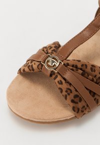 Brown high-heeled sandal with a leopard print fabric upper, a knotted design, and a gold circular accent on a smooth tan footbed.