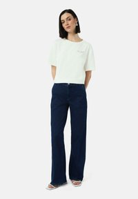 Weißes T-Shirt mit kurzen Ärmeln und Rundhalsausschnitt, kombiniert mit dunkelblauen weiten Jeans. Das Modell trägt minimalistische weiße Sandalen.