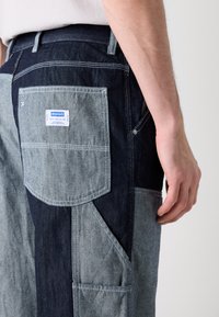 Denim cargo pants featuring a mix of light and dark blue shades, multi-pocket design, and a tag with brand logo on the back pocket.