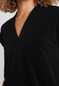 Kvinna med lockigt hår som bär en svart V-ringad topp mot en neutral bakgrund, visande överkropp och nacke.