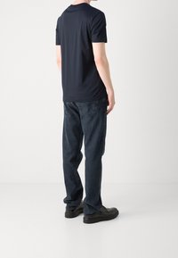 Man som står, iklädd en marinblå kortärmad skjorta, mörkblå jeans och svarta skor, sedd bakifrån mot en neutral bakgrund.