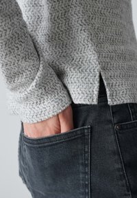 Grauer gemusterter Pullover mit strukturiertem Finish, kombiniert mit schwarzen Jeans mit einer Vordertasche. Der Pullover hat einen seitlichen Schlitz am Saum.