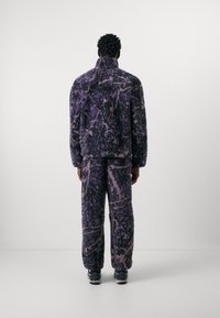 Fleece jacket and matching pants in purple and black abstract pattern, featuring a high collar and relaxed fit, paired with black footwear.