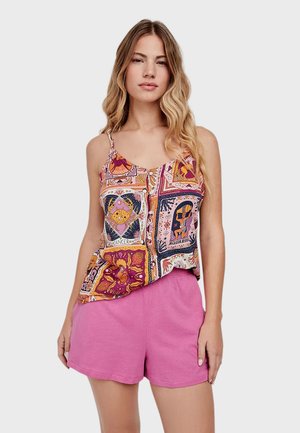 Woman with wavy blonde hair wearing a colorful zodiac print camisole and pink shorts, standing against a plain gray background.