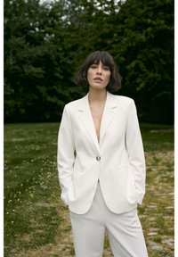 Witte op maat gemaakte blazer met een enkele knoop en notch revers, gecombineerd met bijpassende broeken, tegen een grasachtige achtergrond.
