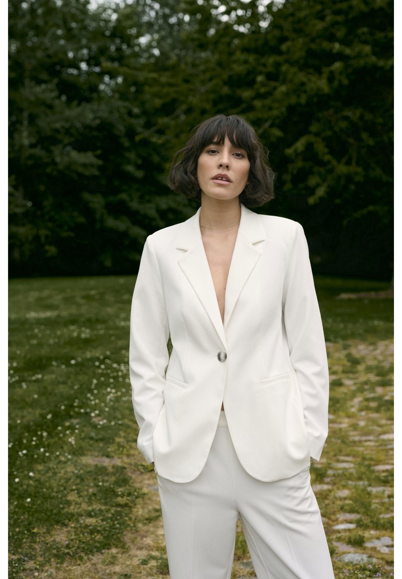 Witte op maat gemaakte blazer met een enkele knoop en notch revers, gecombineerd met bijpassende broeken, tegen een grasachtige achtergrond.