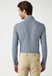 Langarmhemd in blauw met een geometrisch patroon, voorzien van een puntkraag en knoopmanchetten. Gecombineerd met beige broeken.