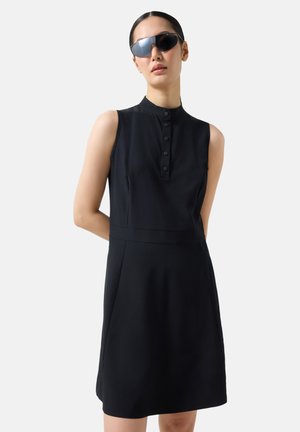 Vrouw met een mouwloze zwarte jurk met een knoopkraag en een zwarte zonnebril, staand met haar handen achter haar rug.