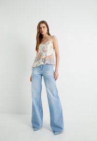Camiseta de tirantes finos en color crema con estampado floral, que presenta un dobladillo de encaje asimétrico, combinada con jeans de pierna ancha en azul claro y tacones de punta.