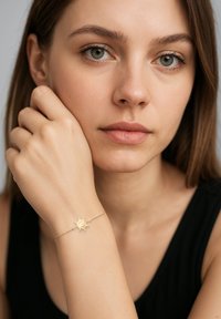 Bracciale d'oro con un ciondolo a forma di sole, caratterizzato da un design testurizzato e una delicata catena. Il portatore ha un'espressione neutra, con pelle chiara e capelli castani.