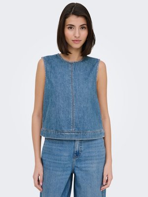 Jeune femme aux cheveux raides et foncés portant un top en denim bleu sans manches et un jean bleu taille haute sur un fond clair uni.