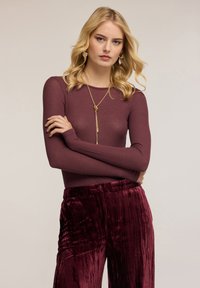 Weinrot langärmliges Oberteil mit gerippter Textur, kombiniert mit samtigen Hosen in passender Farbe. Eine goldene Halskette ergänzt das Outfit.