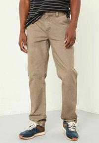 Light brown cotton pants with straight leg design, visible pockets, and a textured finish, paired with navy and brown sneakers.