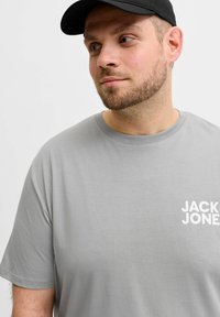 Man with beard wearing black cap and light gray T-shirt with white "JACK & JONES" logo, looking to his left against white background.