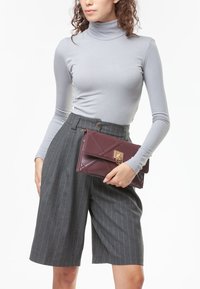 Clutch in pelle bordeaux con design trapuntato e chiusura con accento dorato. Il modello indossa un maglione grigio a collo alto e pantaloni corti grigi a righe.