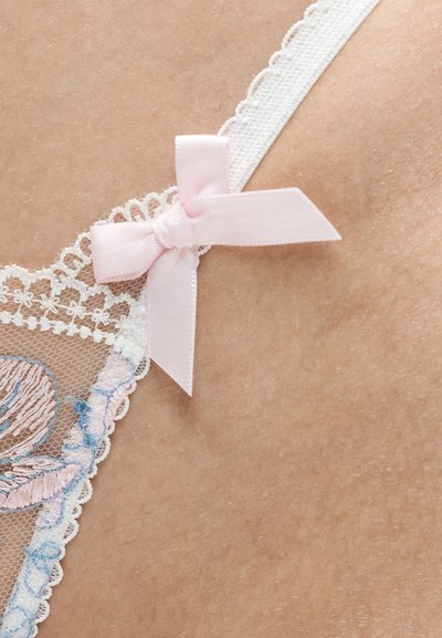 Λεπτή λευκή δαντελένια τιράντα εσώρουχου με ροζ φιόγκο πάνω στο δέρμα, με floral κεντήματα σε μπλε και μπεζ αποχρώσεις.