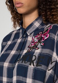 Rutig skjorta i marinblå och rosa med blommig broderi. Har en kragig nacke och stora svarta bokstäver som tillför textur och designintresse.