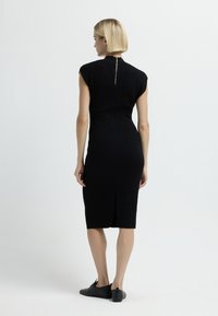 Vestito nero di lunghezza al ginocchio con maniche corte a cappuccio, tessuto texture, chiusura con zip sul retro e uno spacco all'orlo. Indossato con scarpe nere.