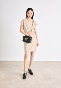 Robe en tricot beige avec un décolleté en V et une taille froncée, associée à un sac bandoulière en cuir noir avec un logo doré. Mules à talons blocs noirs.