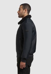 Musta bombere takki, jossa on turkistryyppinen kaulus, sivu-vetoketjullinen taskudetail ja ribatoidut hihansuut. Materiaali näyttää sileältä ja hieman pehmustetulta.