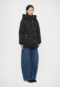 Must kapuutsiga puffer-jope, millel on tõmblukk ja presskinnitused, küljetaskud ning soonikkoes mansetid. Ühestatud laia säärega siniste teksapükstega.