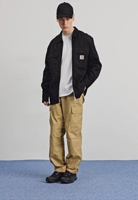 Sort lærredsjakke med to brystlommer, hvid t-shirt under, tan cargo-bukser med flere lommer og sorte sneakers.