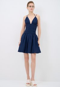 Robe bleu marine avec un décolleté plongeant, un corsage froncé et une jupe évasée. Confectionnée dans un tissu texturé. Associée à des talons compensés beiges.