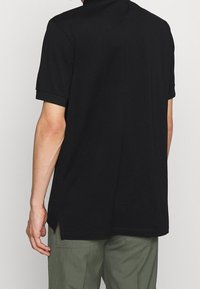 Svart kortärmad polo med krage, tillverkad av mjukt tyg. Har en rak nederkant med sidospaltnar och en ren, minimalistisk design.