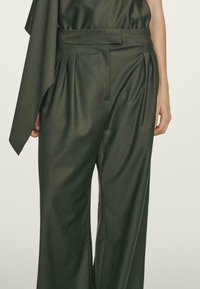 Spodnie o wysokim stanie w ciemnooliwkowym kolorze, szerokie nogawki z plisami z przodu, noszone z dopasowaną ciemnooliwkową bluzką z asymetrycznym rękawem.