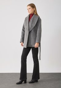 Manteau en laine grise avec grand col replié, fermeture style enveloppe, poches latérales et taille à nouer. Associé à un jean noir évasé et des bottes à talons.