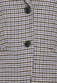 Veste en laine à carreaux noirs et bleus avec deux boutons noirs et deux poches fendues horizontales.