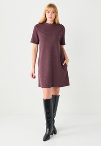 Bordowa sukienka z krótkimi rękawami i wysoko podniesionym dekoltem, z ozdobnym szwem w kształcie litery V oraz bocznymi kieszeniami. Połączona z czarnymi kozakami za kolano.