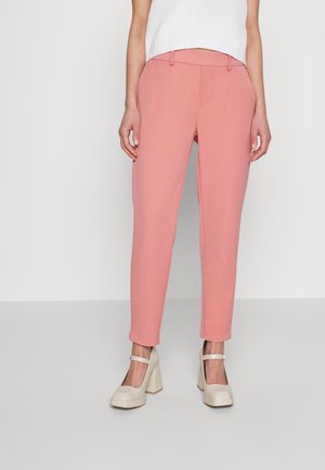 Woman wearing high-waisted, tapered pink trousers paired with white chunky-heeled Mary Jane shoes and a white sleeveless knit top.