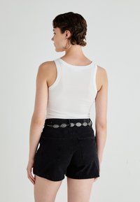 Top din bumbac alb fără mâneci, purtat cu șorturi din denim negre. Șorturile au două buzunare pe spate și o curea decorativă argintie. Texturi netede pe tot parcursul.