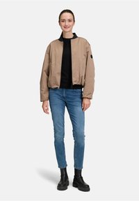 Giacca bomber khaki con polsini e orlo elasticizzati, frontale con zip e colletto nero, abbinata a jeans skinny blu e stivali neri con platform.