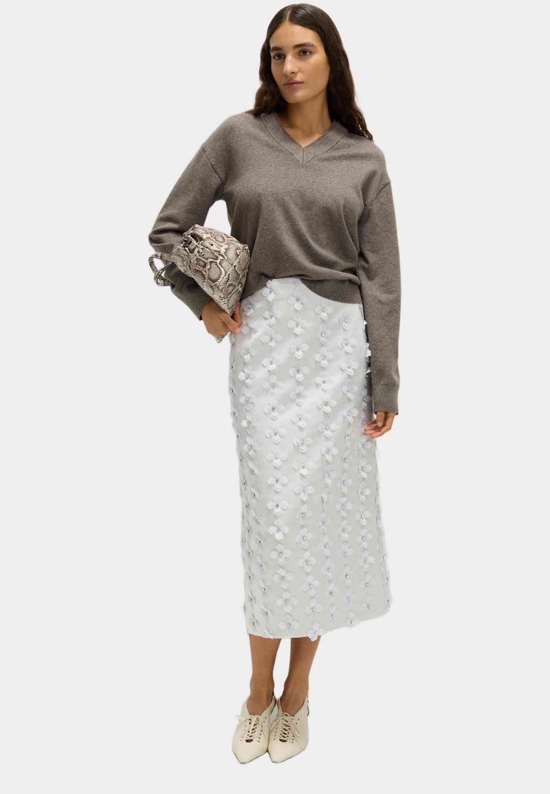 Suéter gris de punto con escote en V, falda midi blanca con adornos circulares, bolso estampado y calzado crema con cordones. Elementos de diseño sencillo.