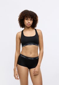 Must musta spordibrassi ja poistepükside komplekt, millel on ruudukujuline kaelus ja elastsed ribad, millel on valge "Calvin Klein" logo. Sile tekstuur.