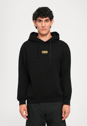 Young man with curly hair wears black hoodie with gold "Versace Jeans Couture" logo, standing against a plain light background.