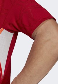 Czerwona koszulka sportowa z teksturowanym geometrycznym wzorem, z białą siateczkową wstawką i pomarańczowymi akcentami po bokach.