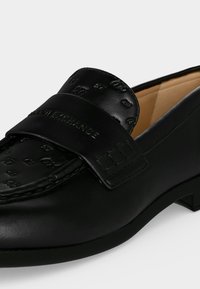 Mocassin en cuir noir avec logo embossé et détail de couture, avec un petit talon et une doublure intérieure couleur beige.