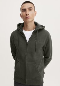Felpa con zip verde scuro realizzata in tessuto morbido, dotata di una tasca frontale, cappuccio con coulisse e un discreto dettaglio del logo sul petto.