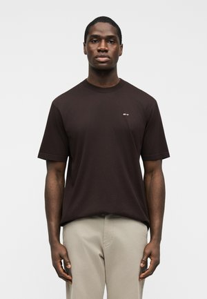 Homme debout portant un t-shirt à manches courtes marron foncé avec un petit texte blanc sur la poitrine et un pantalon beige clair sur un fond blanc.
