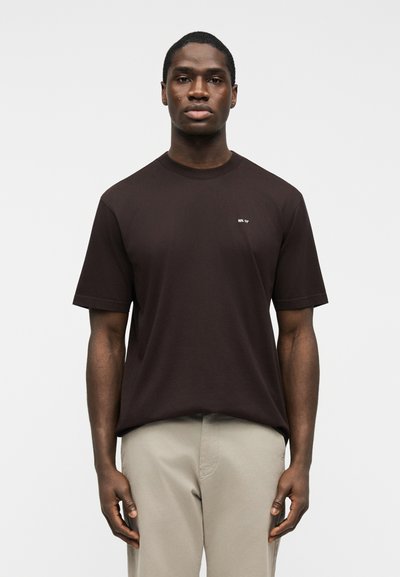 Homme debout portant un t-shirt à manches courtes marron foncé avec un petit texte blanc sur la poitrine et un pantalon beige clair sur un fond blanc.