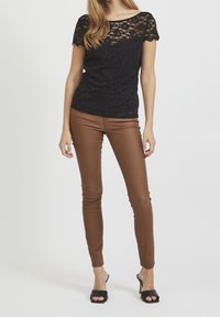 Schwarzes Spitzenoberteil mit kurzen Ärmeln und einem gewellten Ausschnitt, kombiniert mit engen braunen Kunstlederhosen und schwarzen offenen Schuhen mit Absatz.