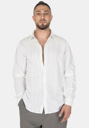 Uomo con barba e capelli legati indossa una camicia bianca a maniche lunghe con fantasia parzialmente sbottonata e pantaloni grigi, in piedi con le mani nelle tasche.