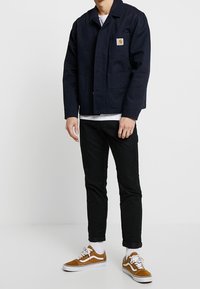 Marineblauwe jas met een knoopsluiting aan de voorkant, twee grote zakken en een logo-patch. Draagt het met zwarte broek en bruine suède sneakers met witte accenten.